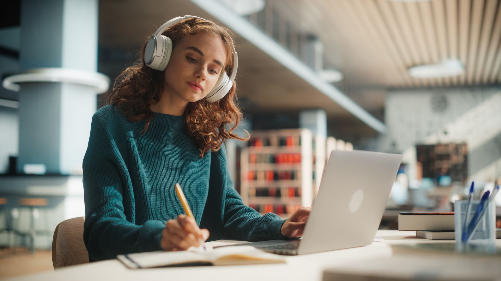 Concentrated Young Female Student Engaged in Academic Research With Laptop and Headphones at Modern Library Desk, Writing Notes and Studying Online.