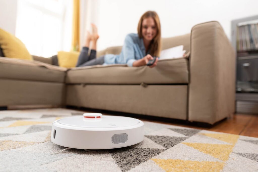 Robotic vacuum cleaner cleaning the room while woman relaxing on sofa. Woman controlling vacuum with remote control.