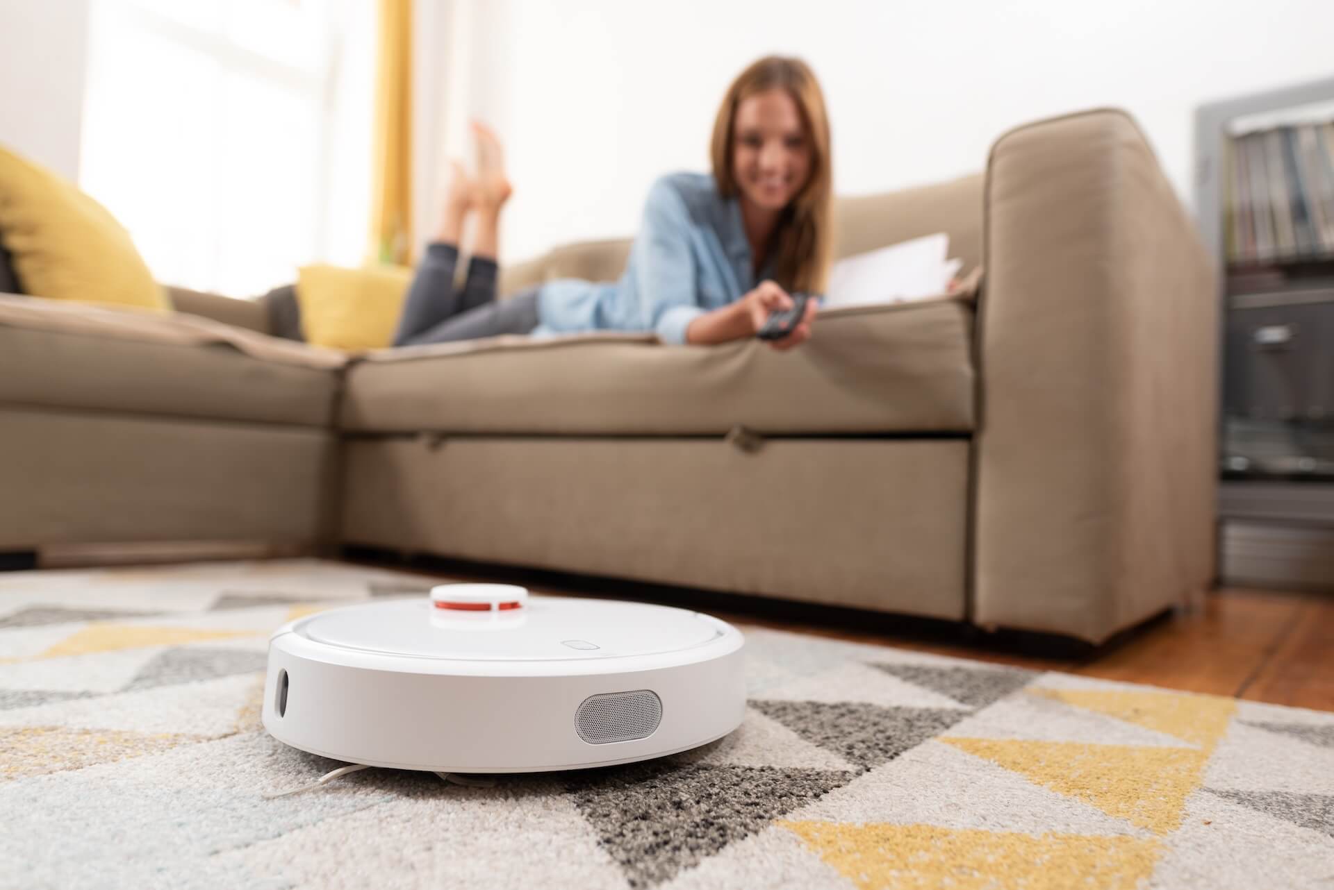 Robotic vacuum cleaner cleaning the room while woman relaxing on sofa. Woman controlling vacuum with remote control.
