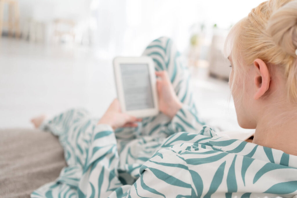 A woman relaxing on a couch in patterned loungewear while reading on an e-reader tablet in a bright living room