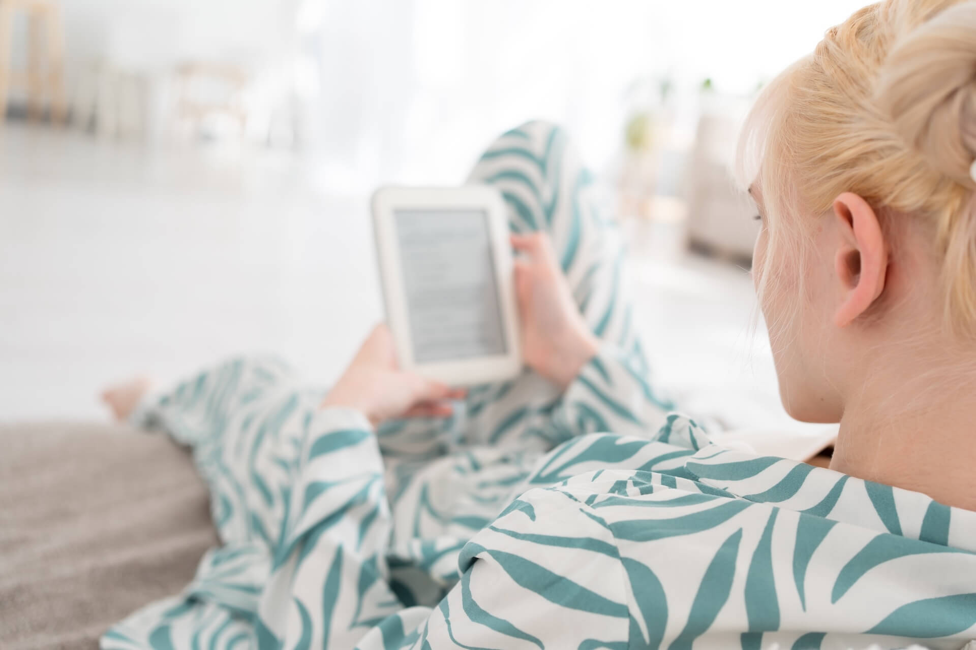 A woman relaxing on a couch in patterned loungewear while reading on an e-reader tablet in a bright living room