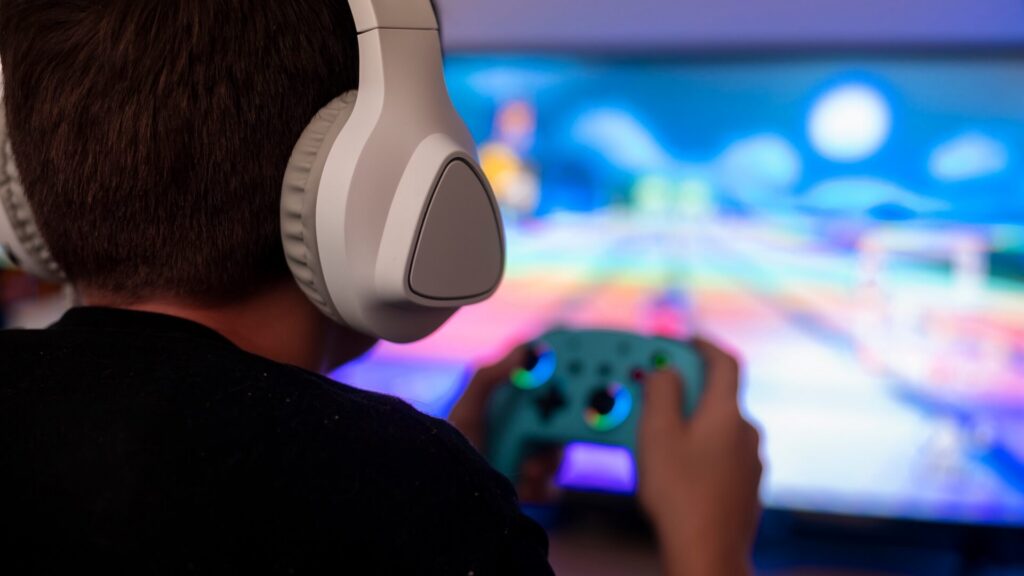 Close-up from behind of a child wearing white headphones, holding a game controller while playing a brightly colored video game on a screen
