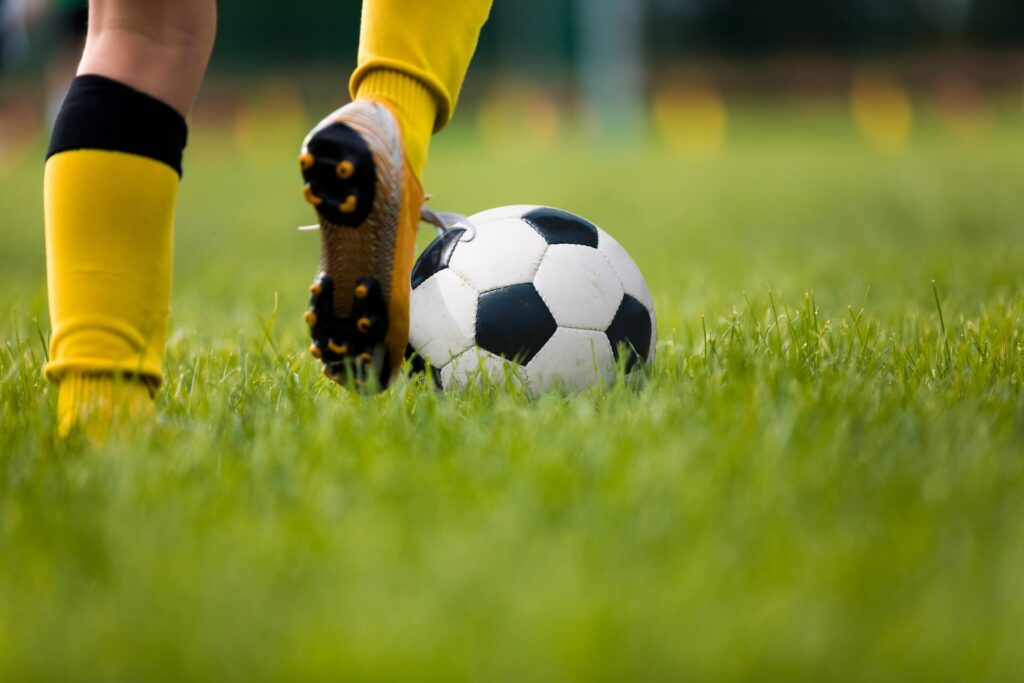 A close-up of a football player wearing yellow socks and cleats, running and stepping toward a black-and-white football on green grass