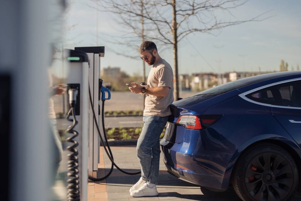 Man using his phone while charging an electric car at a public charging station.