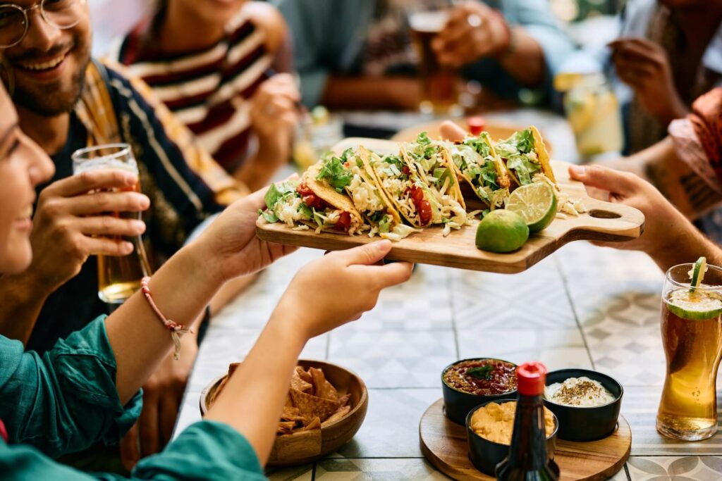 People at a restaurant table sharing a wooden board of tacos topped with lettuce, cheese, and salsa, with lime wedges on the side, while drinks and dips sit on the table and friends chat in the background.