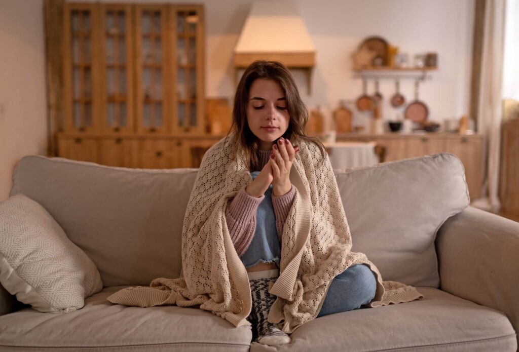 Woman wrapped in a blanket sitting on a couch, holding her hands together in a cozy living room.