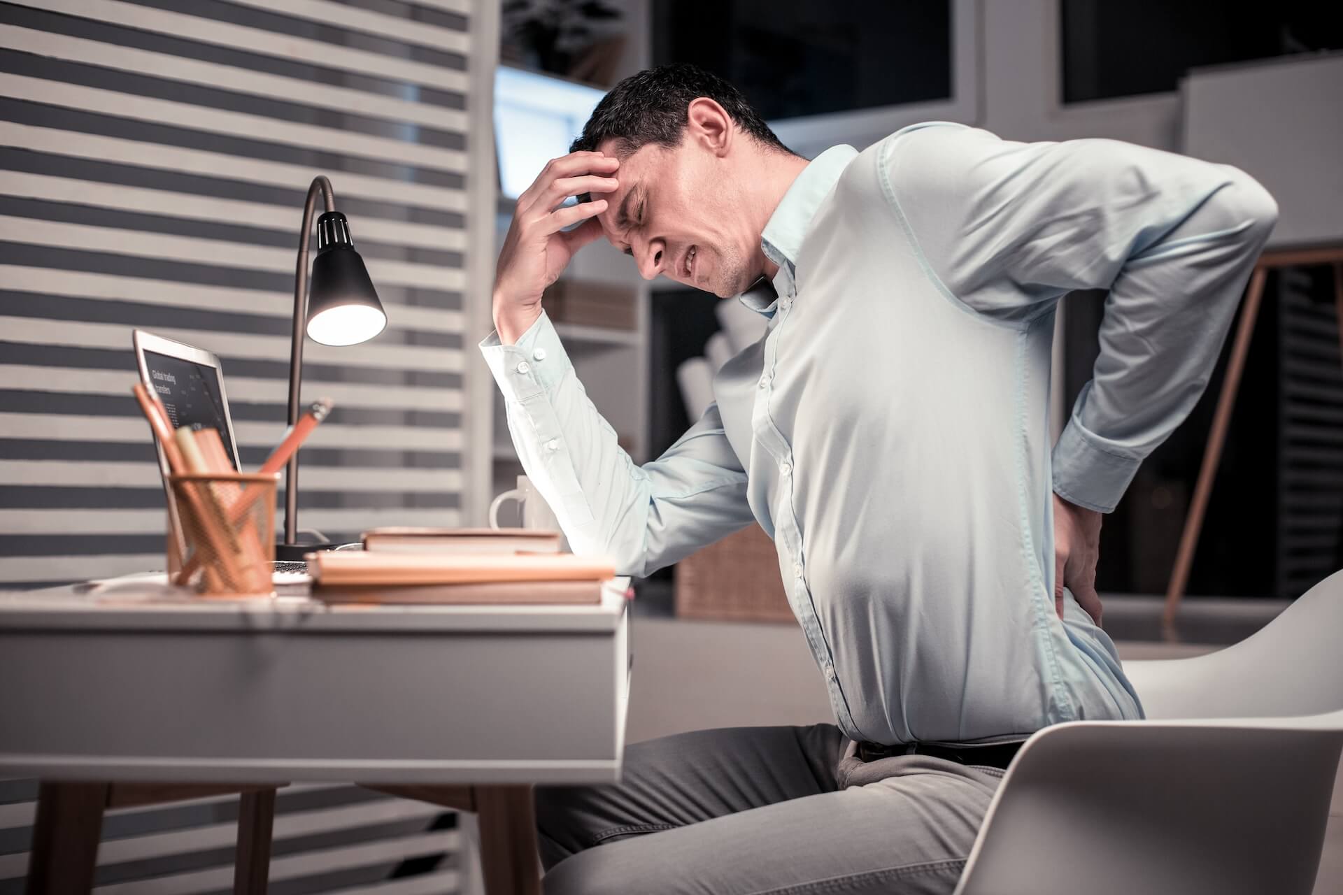 Man sitting at a desk holding his lower back in pain while working on a laptop.