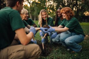 **Alt text:** A diverse group of volunteers wearing green shirts and gardening gloves kneel together in a park, smiling as they plant a young tree in the soil during a community environmental activity. 🌱