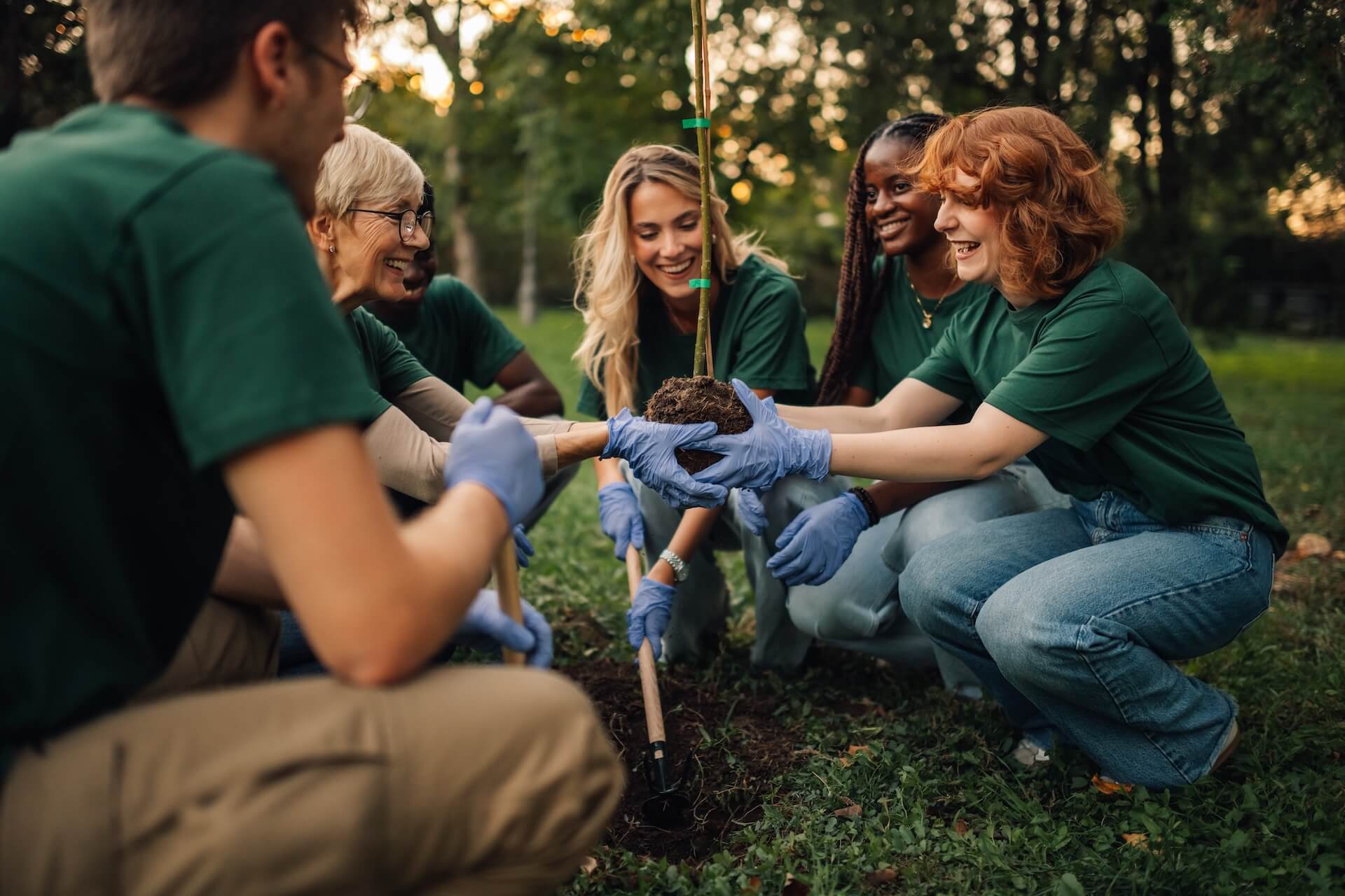 **Alt text:** A diverse group of volunteers wearing green shirts and gardening gloves kneel together in a park, smiling as they plant a young tree in the soil during a community environmental activity. 🌱