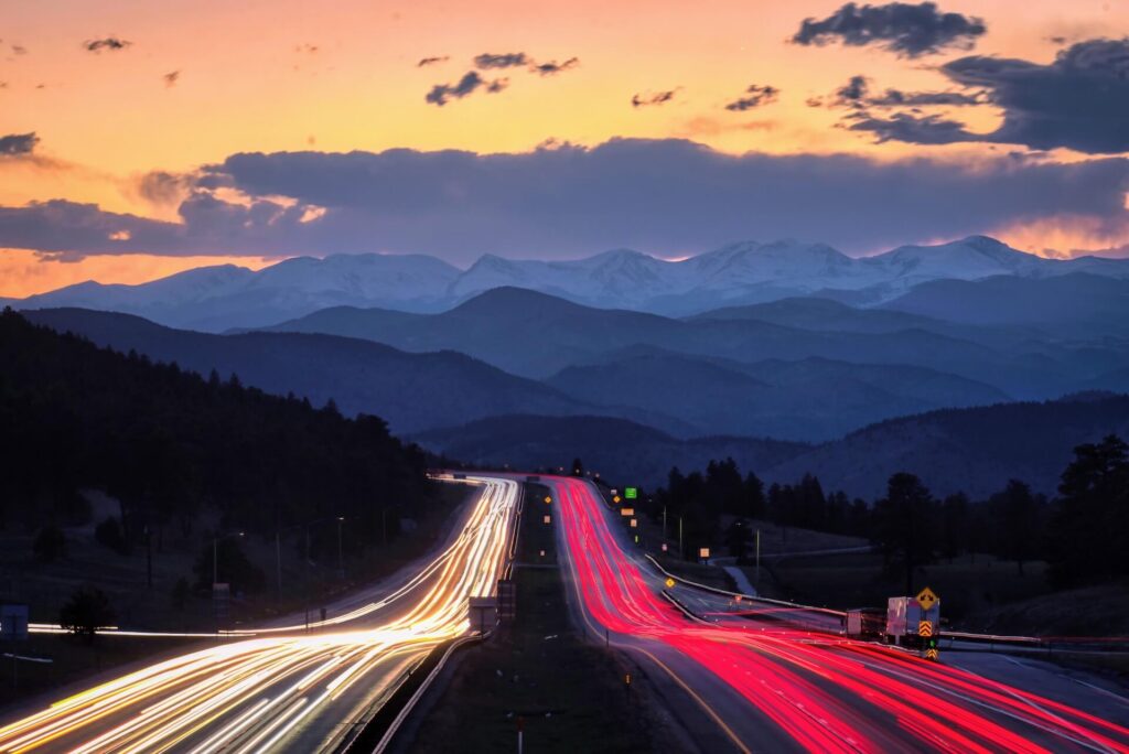 Highway light trails leading toward snow-capped mountains at sunset.