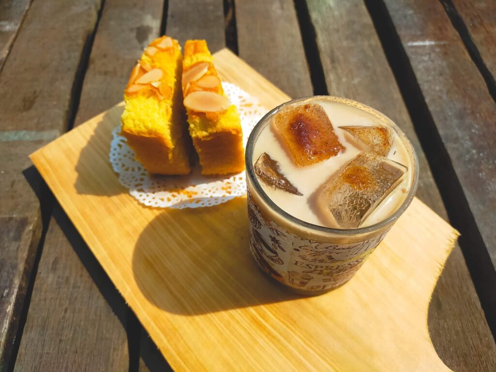 Iced coffee with ice cubes and two slices of almond-topped cake on a wooden board outdoors.