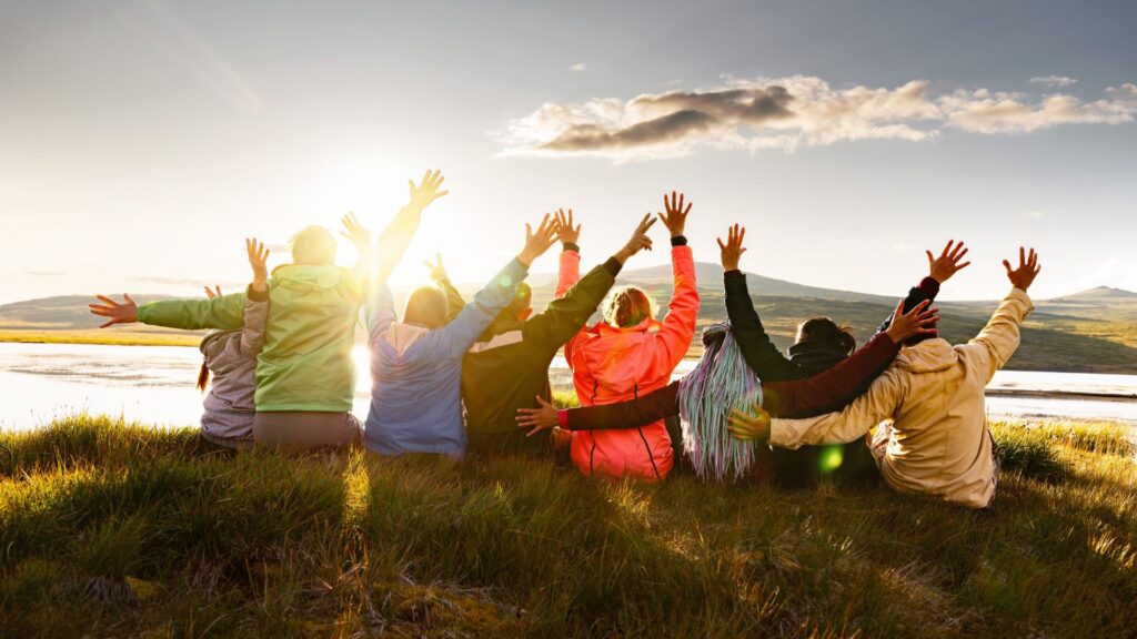 Large group of happy friends sitting by mountain lake with raised arms at sunset, celebrating travel and freedom