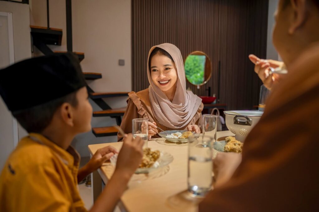 A smiling woman wearing a light-colored hijab shares a meal with family members at home while celebrating Idul Adha together.