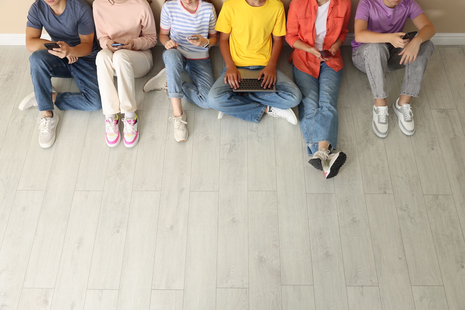 Young people sitting on the floor using smartphones and a laptop, with empty floor space in the foreground.