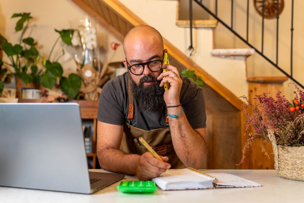 Man on phone writing notes beside a laptop and calculator.