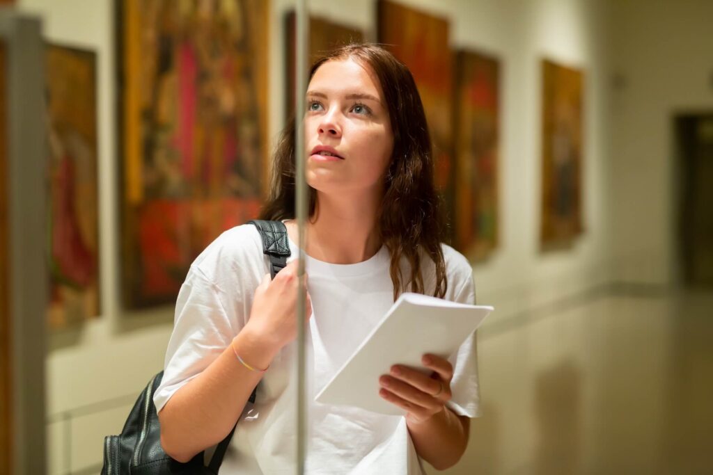 Woman with a backpack observing paintings in an art gallery.