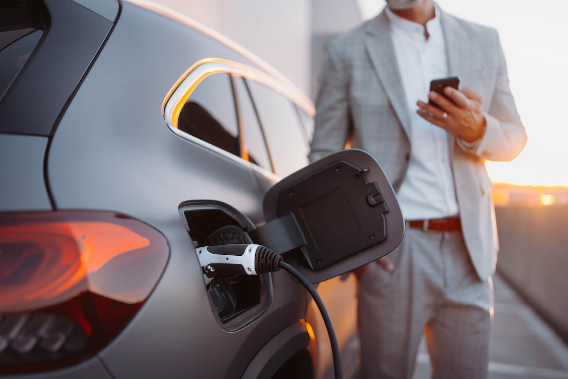 Electric vehicle charging while a person checks their phone nearby at sunset.
