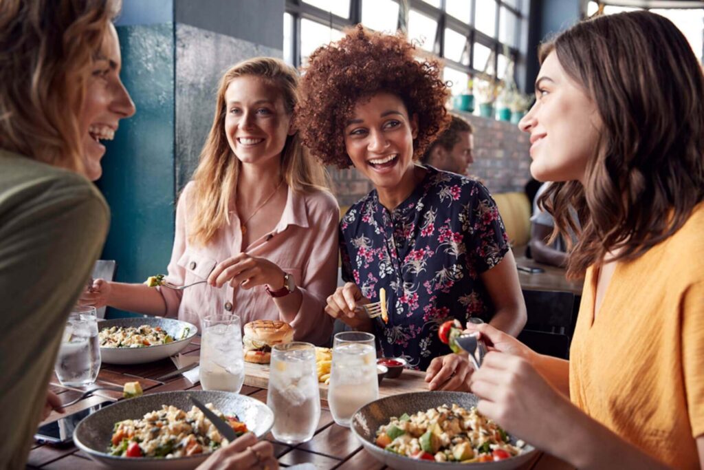 **Alt text:** Four women sitting at a restaurant table, smiling and chatting while eating salads and other dishes together in a bright, casual dining space. Glasses of water and plates of food are on the wooden table, and large windows let in natural light behind them