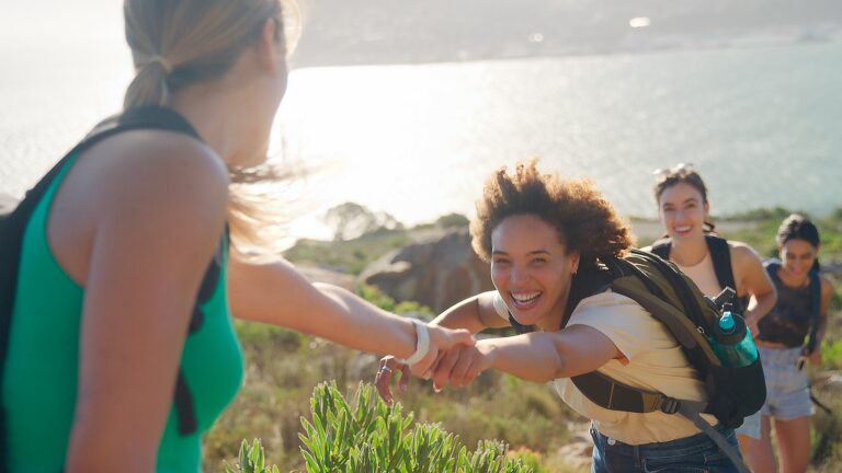 Group Of Female Friends With Backpacks Helping Each Other On Hike In Countryside On Coastal Path