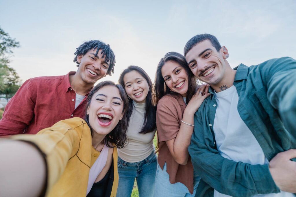 Five young adults smiling and taking a close-up selfie together outdoors on a sunny day, with trees and open sky in the background.