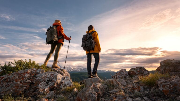 Two girl hikers with backpack stands on top of mountain and look at beautiful sunset sky, back view