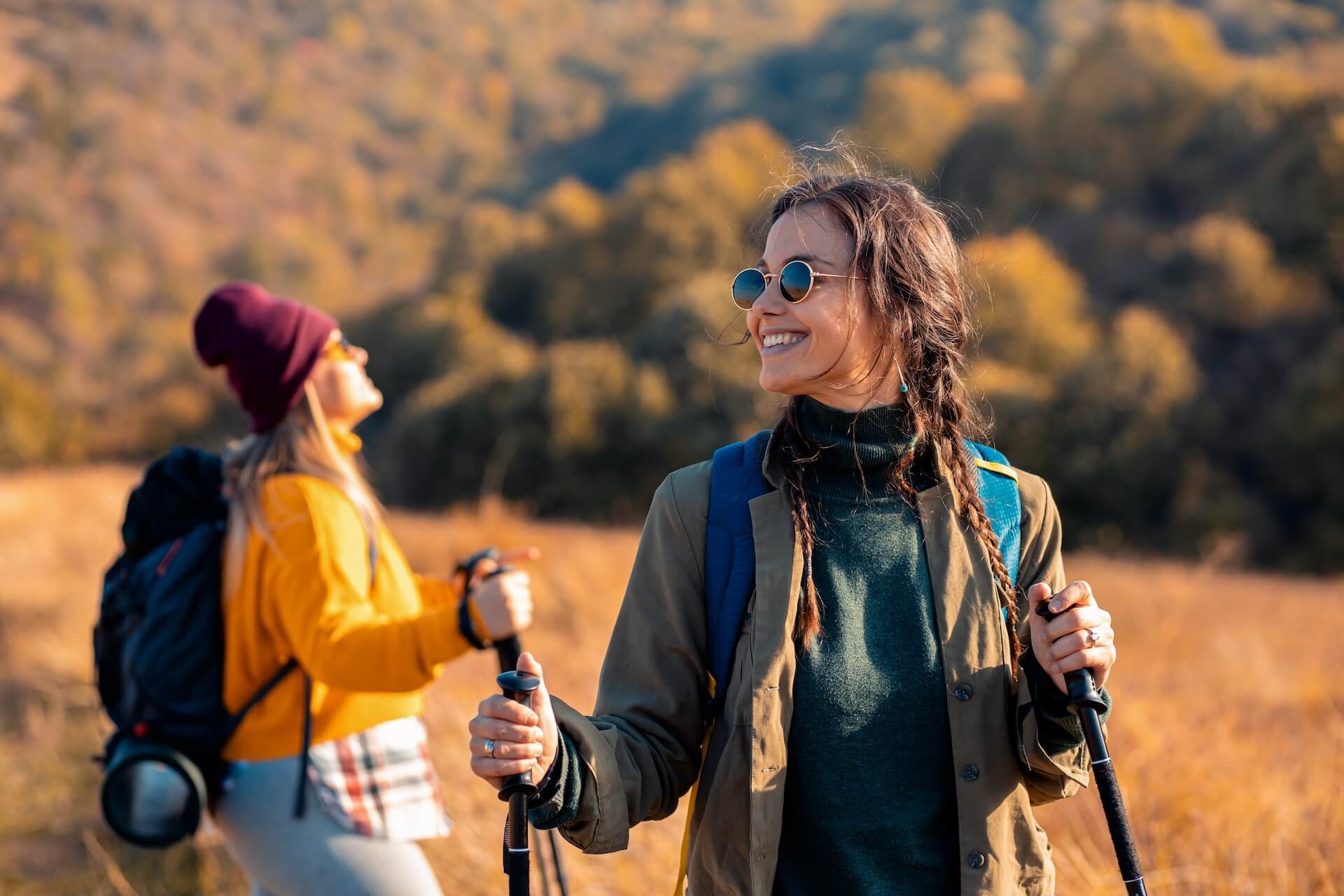 Two women hiking on a sunny autumn day, smiling and using trekking poles, with golden hills and trees in the background