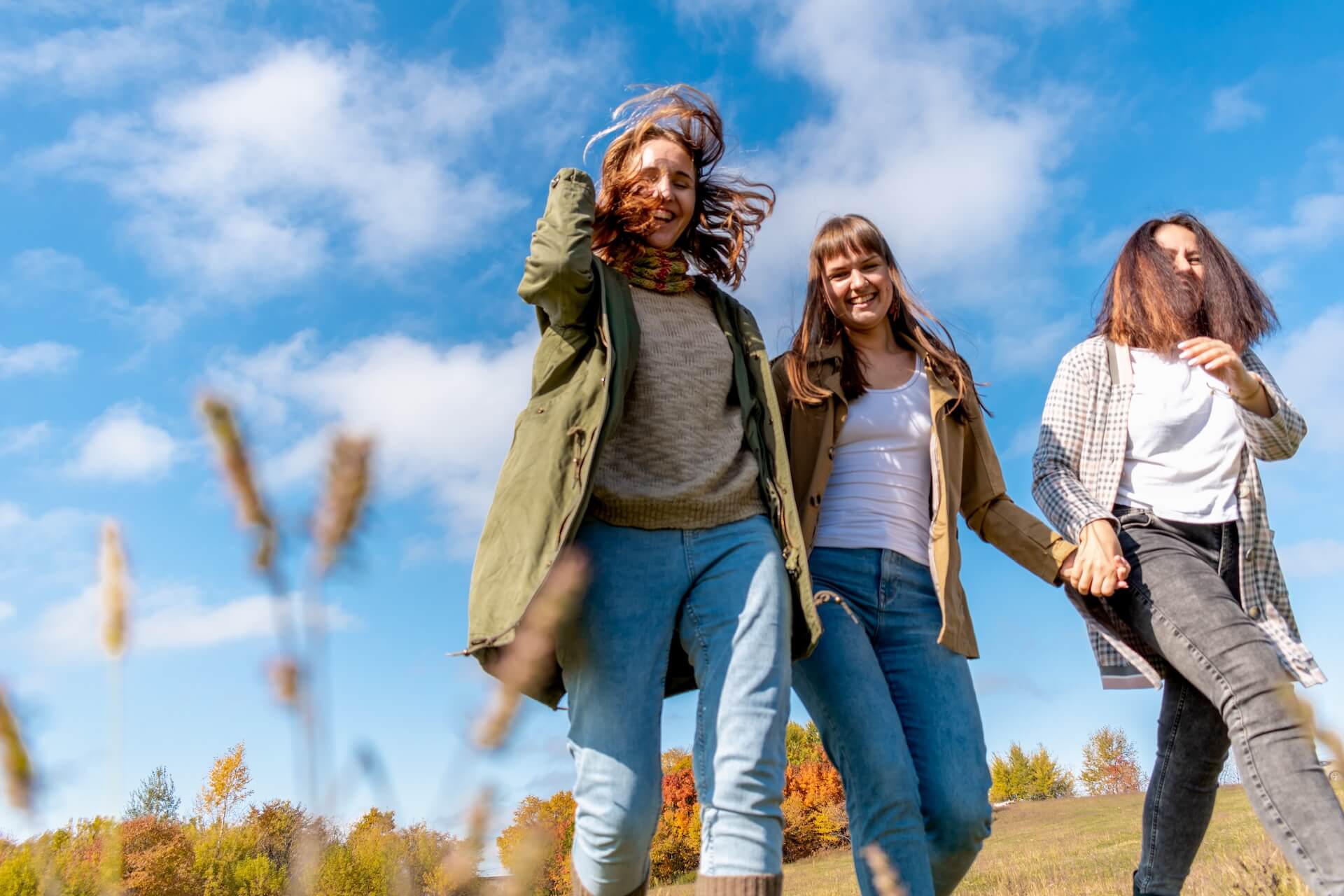 Three friends walking outdoors in a grassy field under a bright blue sky, smiling and holding hands, with autumn-colored trees in the background.
