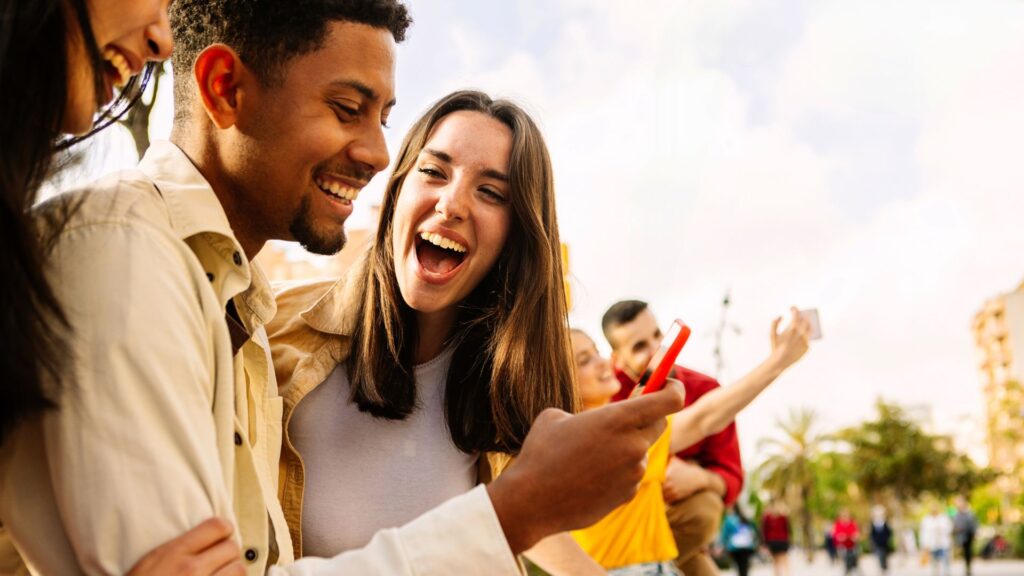 Young adult friends using mobile phone together outside. Gen Z people generation having fun watching social media content on cell. Youth and technology concept