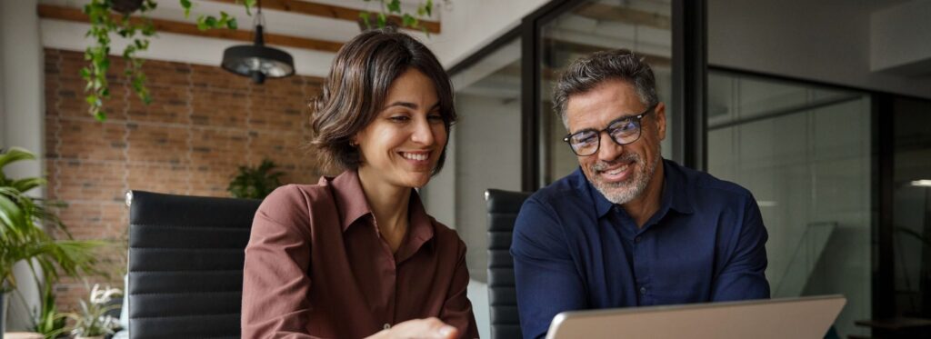 Business team of two executives working together using laptop in office. Happy young business woman manager showing project presentation to mature man client, investor or partner at work meeting.