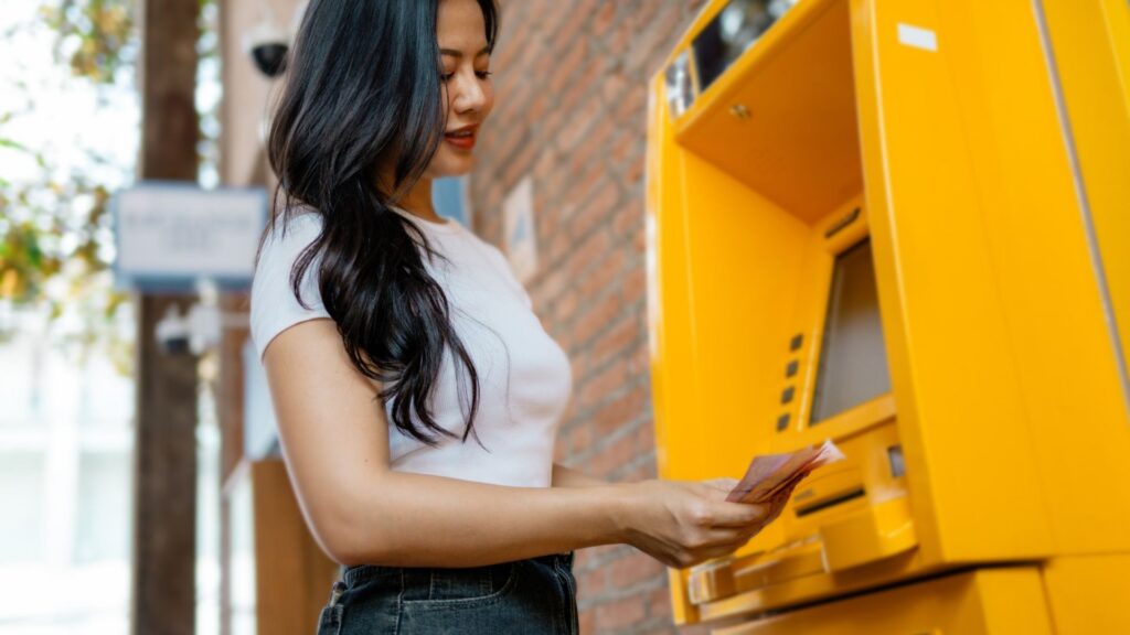 Asian woman withdrawing money from a yellow automated teller machine located in a city