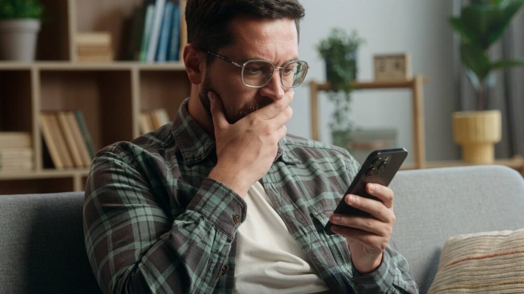 Caucasian adult man male guy at home living room nervous looking at mobile phone bad news amazed shocked close mouth with hand message from bank stressed smartphone notification online error scam
