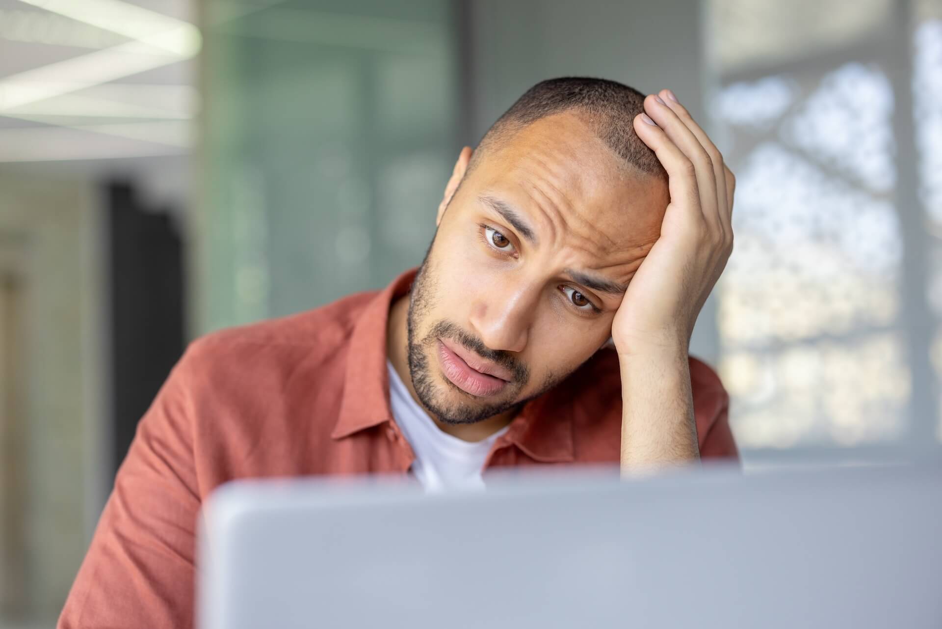 A man looking stressed while sitting at a laptop, resting his head on his hand as he stares at the screen.