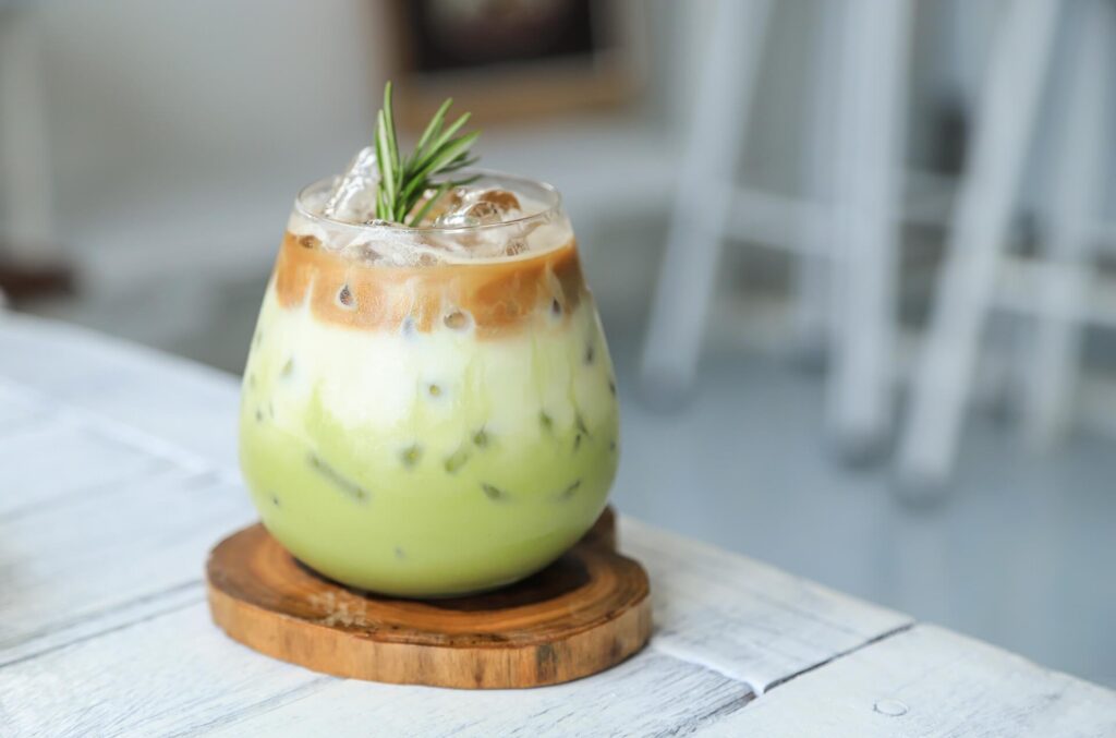 Iced layered matcha latte in a glass, topped with coffee and a sprig of rosemary, sitting on a wooden coaster.
