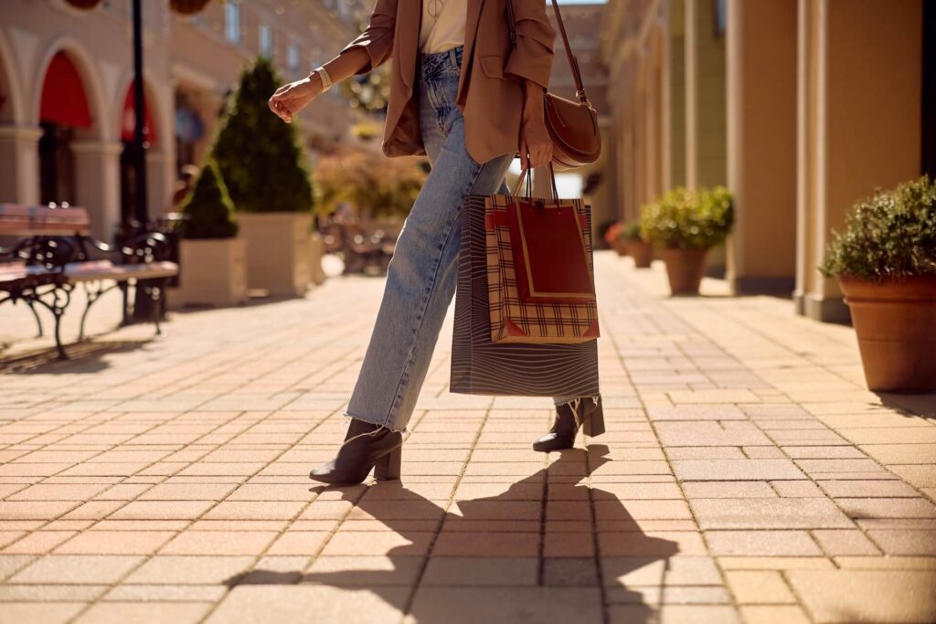 **Alt text:** Person carrying shopping bags while walking through a sunlit outdoor shopping area.