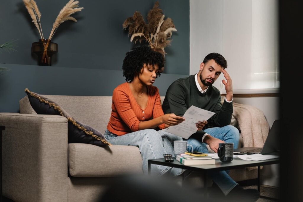 A couple sitting on a couch reviewing bills, looking concerned about their finances.