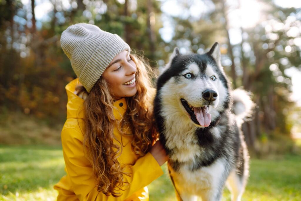 A smiling person in a yellow raincoat and knit beanie kneels beside a husky in a sunlit forest.