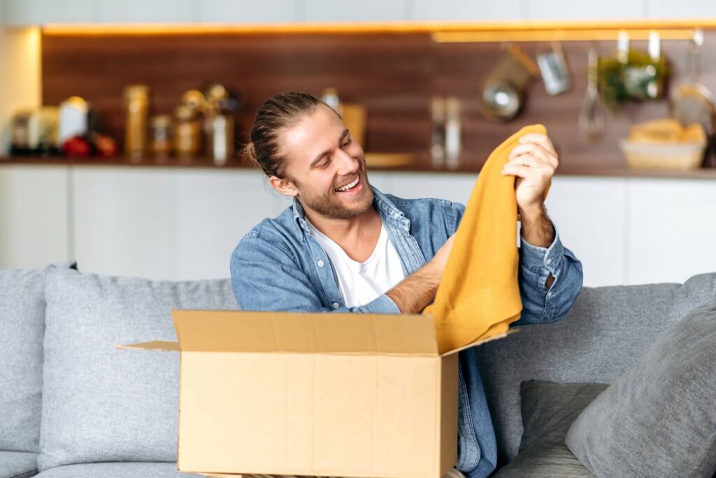 A satisfied smiling guy unpacks his long awaited package, examines the clothes that he bought online