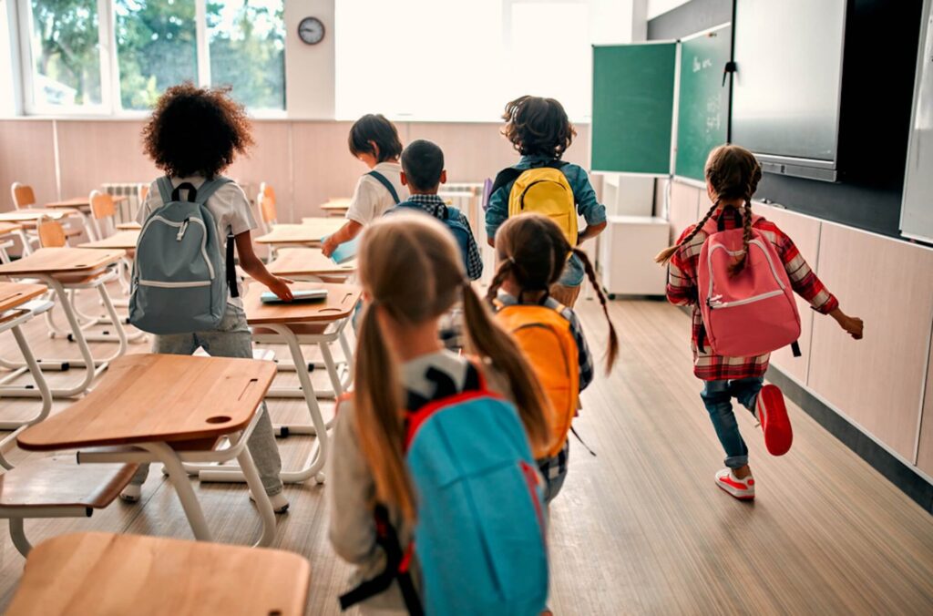 Schoolchildren with colorful backpacks walking into a bright classroom at the start of the school day.