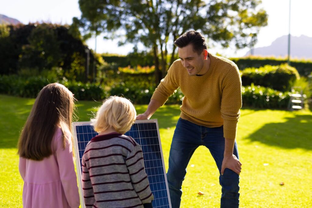 A father in a sunny backyard shows two children a solar panel, smiling as he explains how it works and how it can help save energy