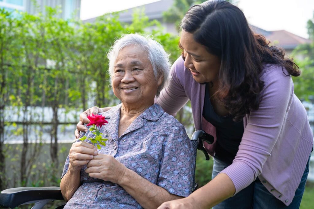 Caregiver daughter hug and help Asian senior or elderly old lady woman holding red rose on wheelchair in park.