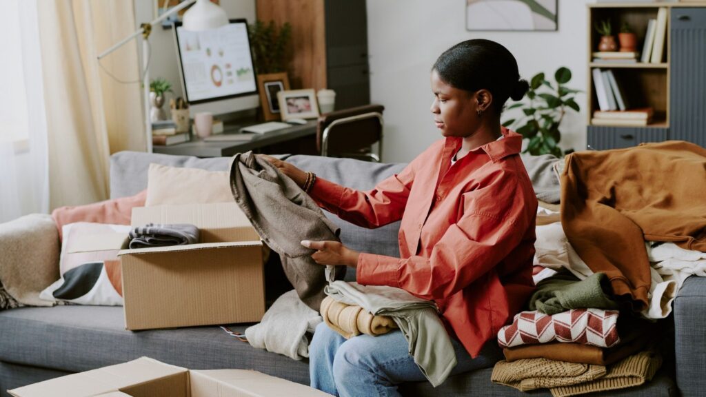 Black young adult woman sorting folded clothes into boxes at home