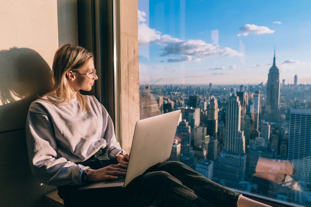 Woman with laptop sitting next to window in city