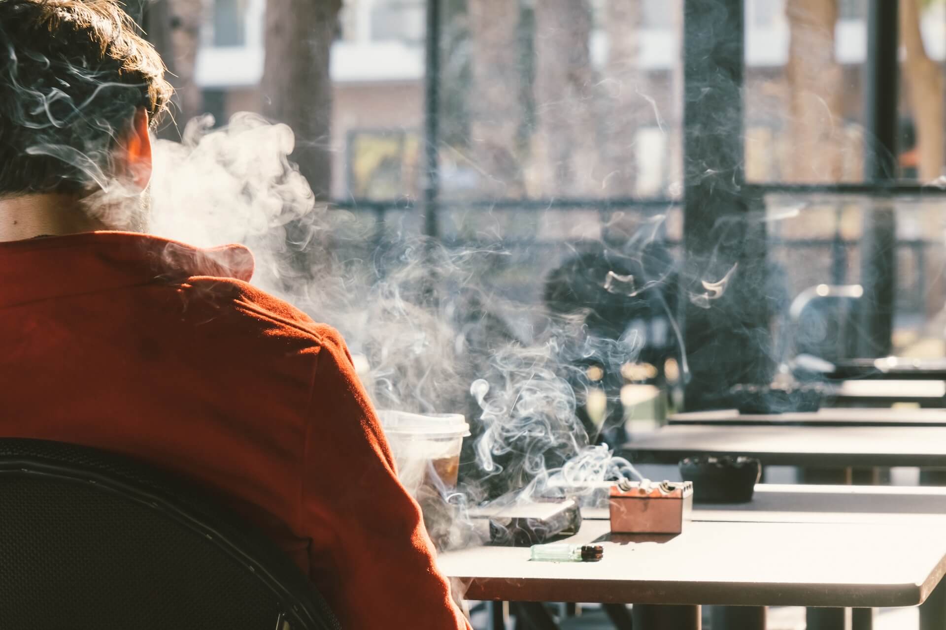 Person in a red jacket smoking at a table with a cigarette and ashtray.
